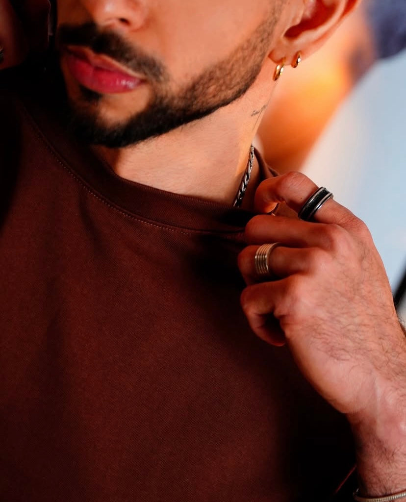 Close-up of a person with a trimmed beard wearing hoop earrings, rings, and a brown Camiseta Tela Pesada 230gr, lightly pulling at the collar. Only the lower face, hand, and upper chest are visible; entrega inmediata disponible.