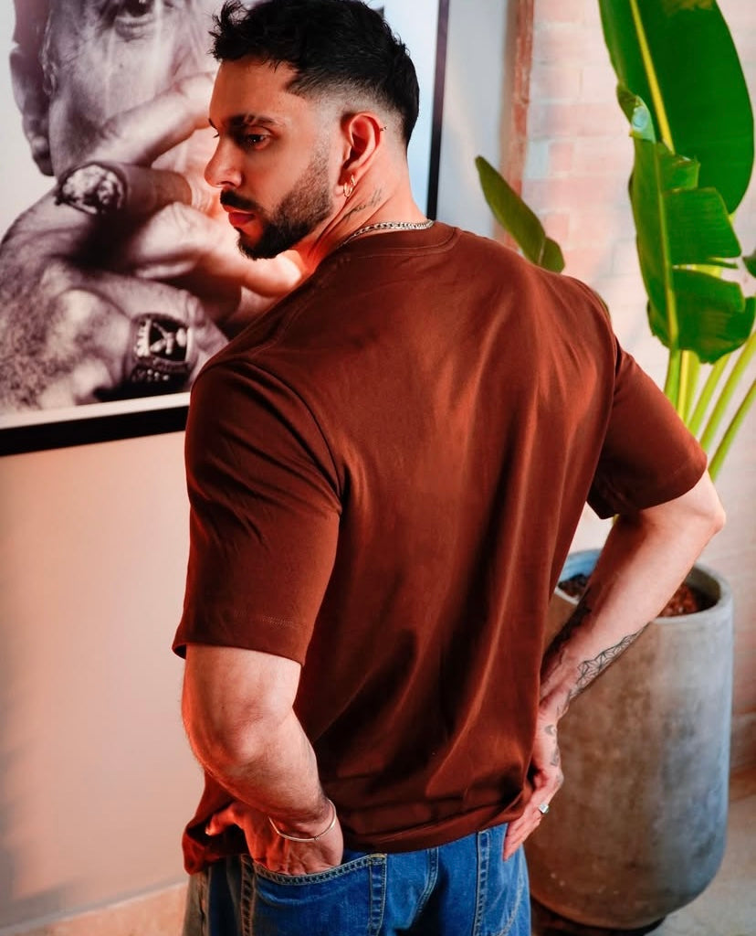 Un hombre de cabello corto oscuro y barba aparece de espaldas, usando la Camiseta Tela Pesada 230gr en color marrón y jeans azules. Está junto a una planta grande; fabricamos bajo pedido con entrega en 4-5 días.