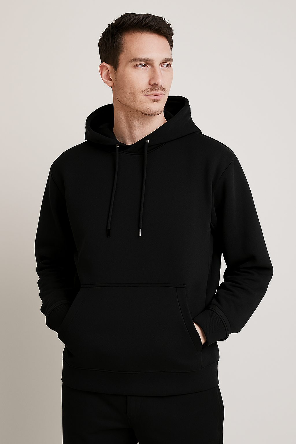 A man models the Hoodie Unisex—plain black with a front pocket and drawstrings—against a light beige background, looking slightly left with hands in pockets. Este modelo está disponible para entrega inmediata.