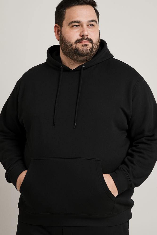 A bearded man wearing a black Hoodie Unisex XL-XXL-XXXL stands with his hands in the pocket against a plain light background, looking slightly to the side. Sudaderas como ésta se fabrican bajo pedido y con entrega inmediata.